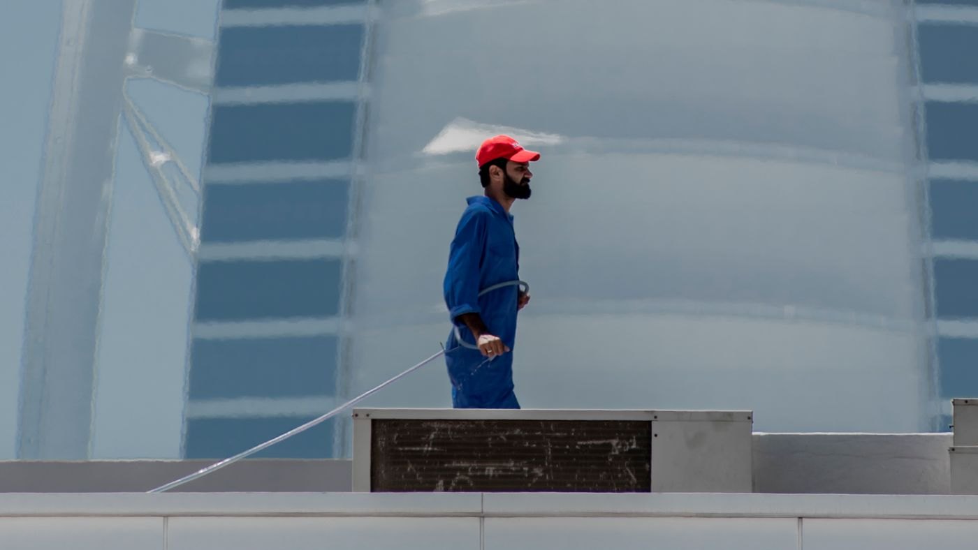 Worker in blue coveralls and a cap on a rooftop beside a plant enclosure, near the roof edge, with the Burj Al Arab behind