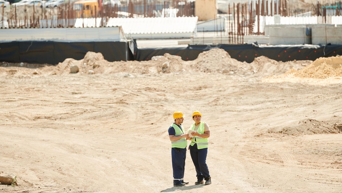 Two workers in hard hats and high-visibility vests standing on a large open construction site of bare earth in bright sun, with no shade structure anywhere nearby