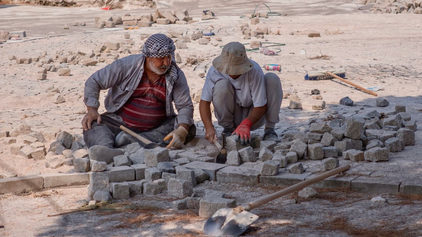 Two road workers laying paving stones on bare ground in full sun, one in a headscarf and one in a wide-brimmed hat, with no shade anywhere in the frame